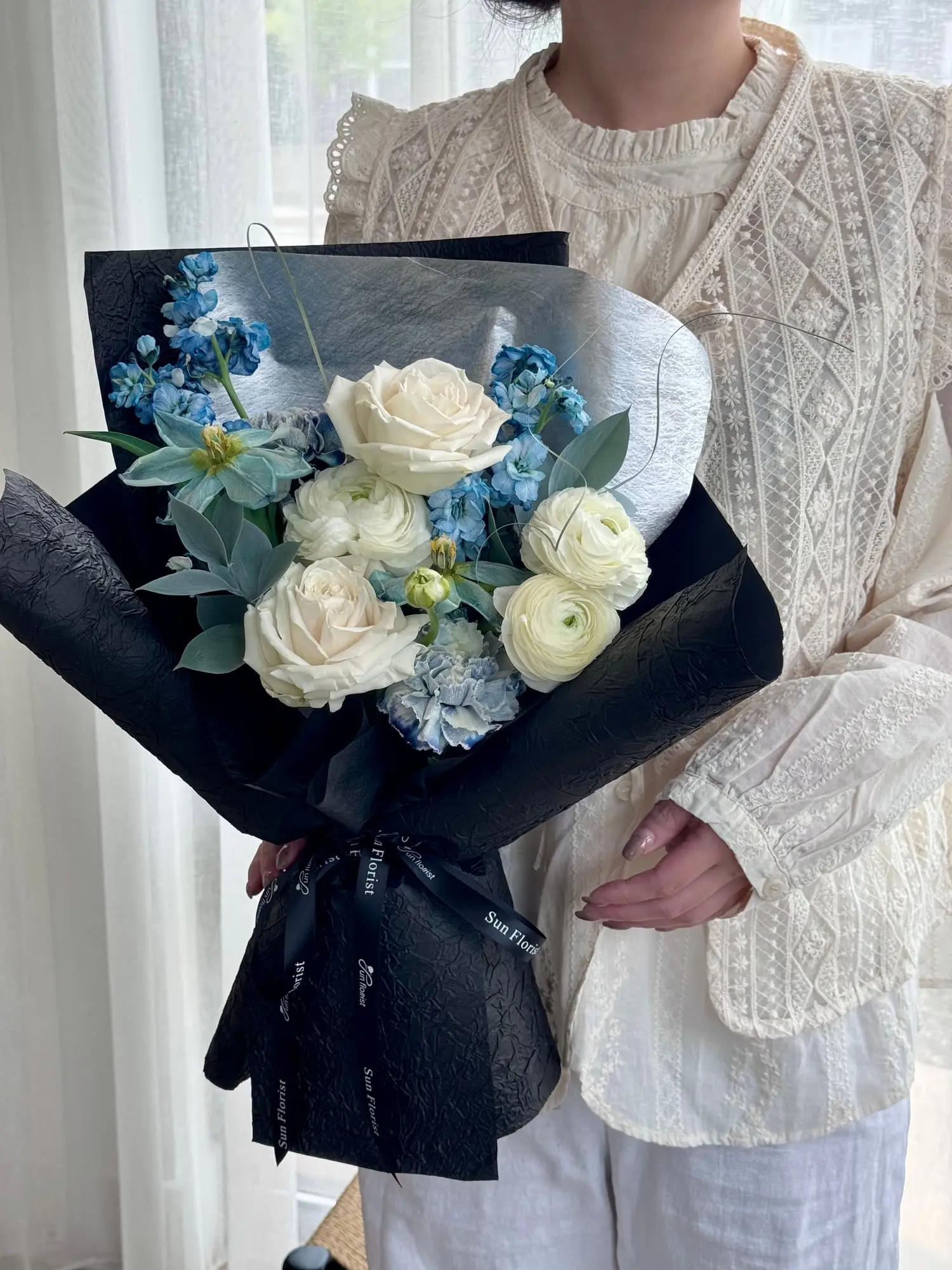 Cool tones, timeless elegance. A modern bouquet of white roses and icy blues, wrapped in black for a touch of mystery.