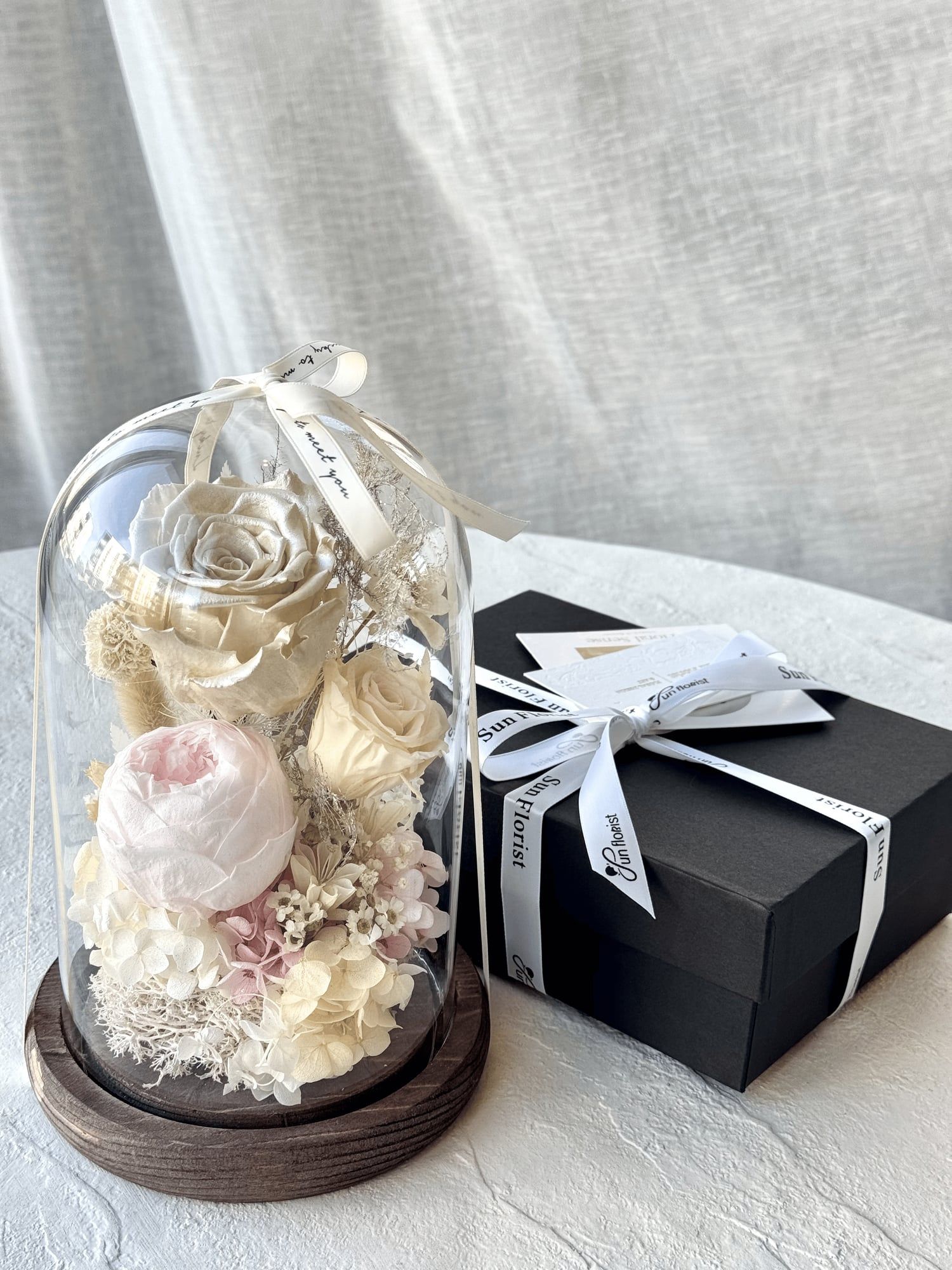 A timeless display of elegance, this preserved flower dome showcases delicate ivory and blush Peony, soft hydrangeas, and pearl-white rose, carefully arranged under a crystal-clear