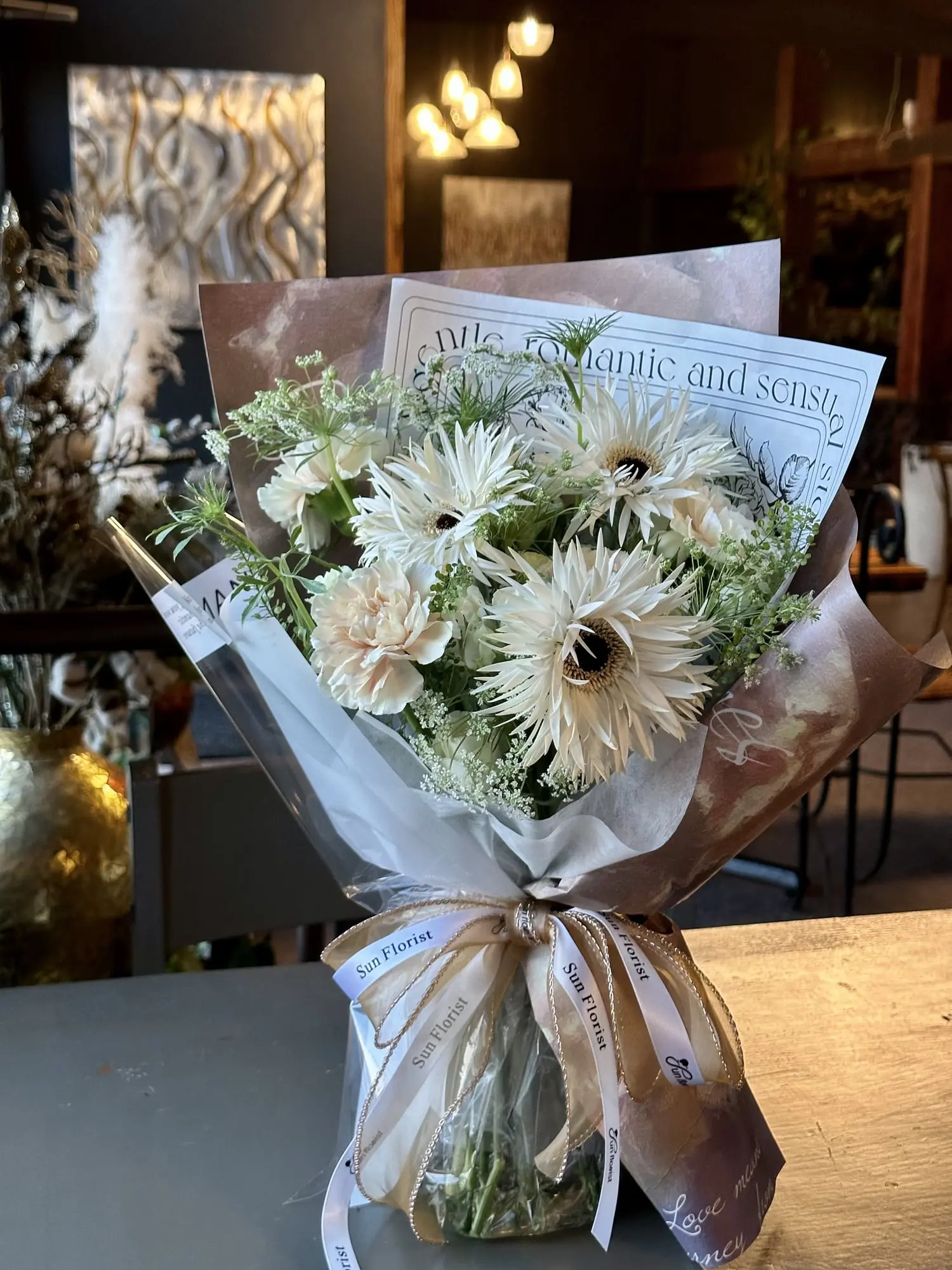 This charming bouquet captures the soft glow of a golden evening with its ivory gerbera daisies, creamy carnations, and delicate greens.
